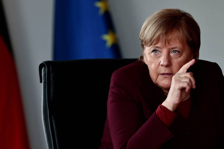 Merkel aponta o dedo esquerdo para cima, de roupa cor de vinho, sentada a uma escrivaninha em frente a uma bandeira azul com estrelas douradas, da UE