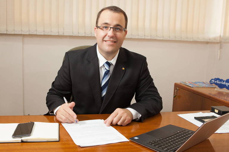 homem branco, de óculos, e terno escuro, sorri, sentado a uma mesa de escritório, com uma caneta na mão