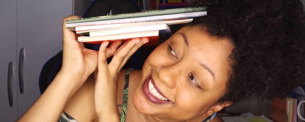 Uma mulher segura com as mãos um pilha de livros e sorri 