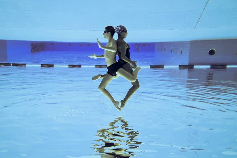 Atletas fazem movimento com os corpos sincronizados, de costas um para o outro, embaixo da água 