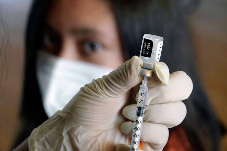Garota de cabelos pretos lisos olha para mão de luva branca que suga vacina de frasco, com seringa