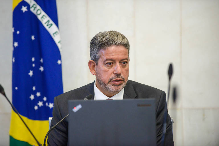 Deputado sentado em frente a tela de computador usando terno e gravata e com bandeira brasileira ao fundo