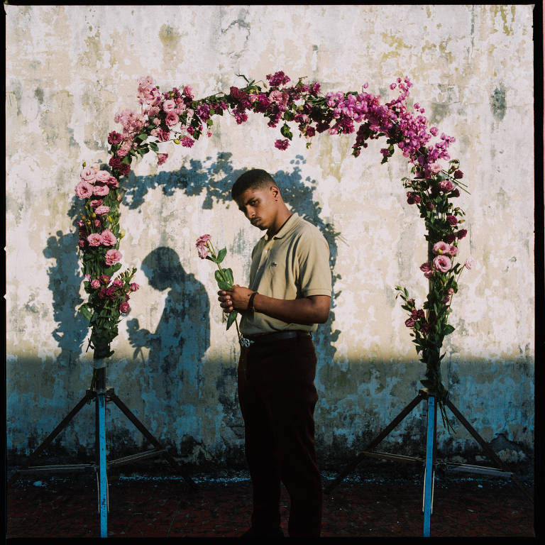 Um homem negro de bigode está em pé, virado de lado, segurando uma flor; ele usa camisa polo bege e calça marrom; atrás dele há um arco coberto de flores