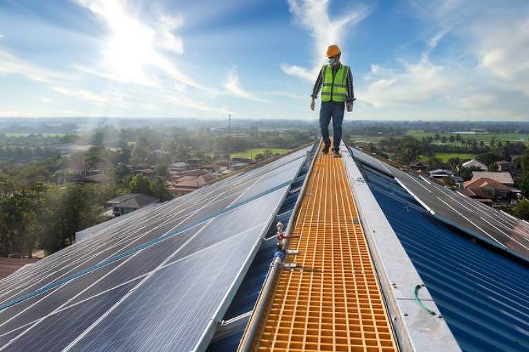 Homem de máscara, capacete e colete fluorescente caminha sobre um telhado com placas metálicas de captação de energia solar