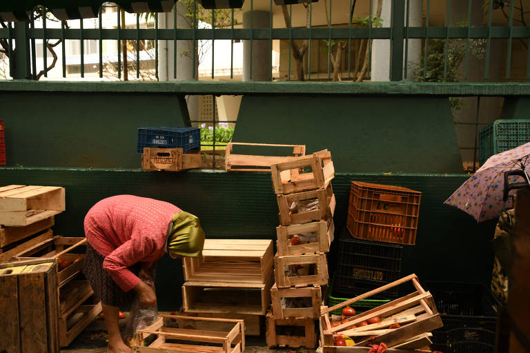 Uma mulher está inclinada, organizando caixas de madeira e plástico em um ambiente de mercado ao ar livre. Há algumas frutas visíveis espalhadas pelo chão, sugerindo que ela está recolhendo essas frutas que seriam desperdiçadas