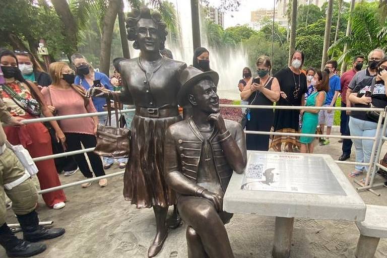 Estátuas do ator Paulo Gustavo e sua personagem Dona Hermínia no centro de um parque, cercado por várias pessoas