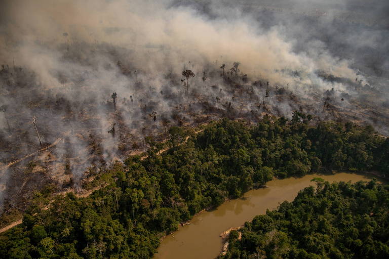 Imagem aérea mostra área de floresta densa sendo queimada. No canto inferior direito, há um rio de águas barrentas