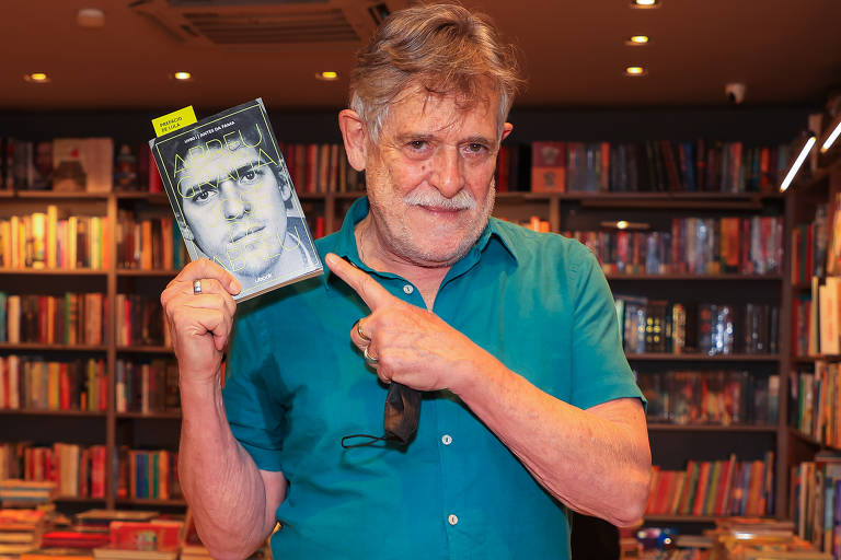 O ator José de Abreu, um homem branco de cabelos e bigode grisalhos, está em pé, de camisa azul, segurando um livro com a capa que leva sua foto
