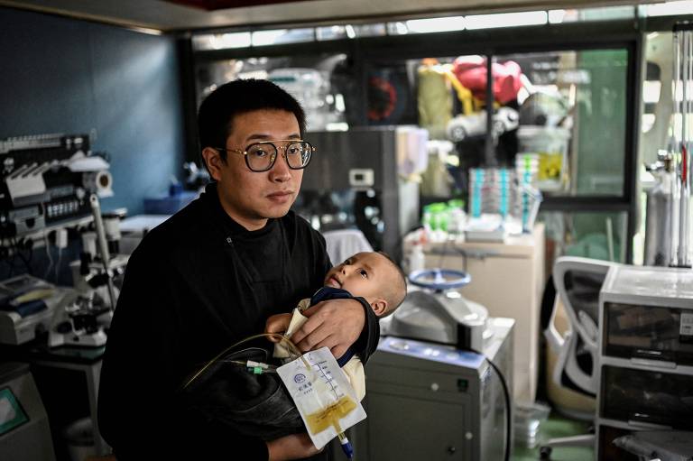 Homem segura bebê no colo em meio à maquinário laboratorial 
