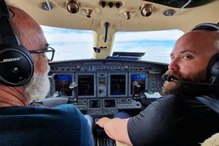 Dentro de uma aeronave, dois homens aparecem de costas na cabine de controle