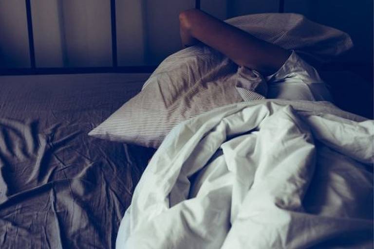 Pessoa deitada em uma cama levao travesseiro para a frente do rosto