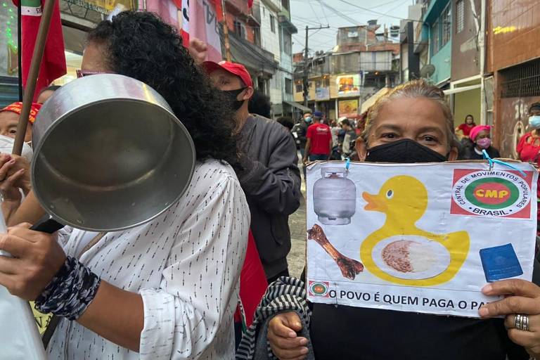 Moradores de Heliópolis fazem Marcha da Panela Vazia. Na foto, uma mulher bate em uma panela e outra segura um cartaz em que se lê "O povo é quem paga o pato".
