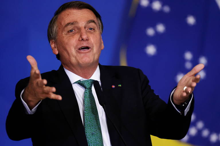 Bolsonaro, um homem branco de cabeços castanhos grisalhos, vestindo terno escuro e com os braços abertos na frente do corpo, gesticulando enquanto fala. Ao fundo dele, uma bandeira do Brasil