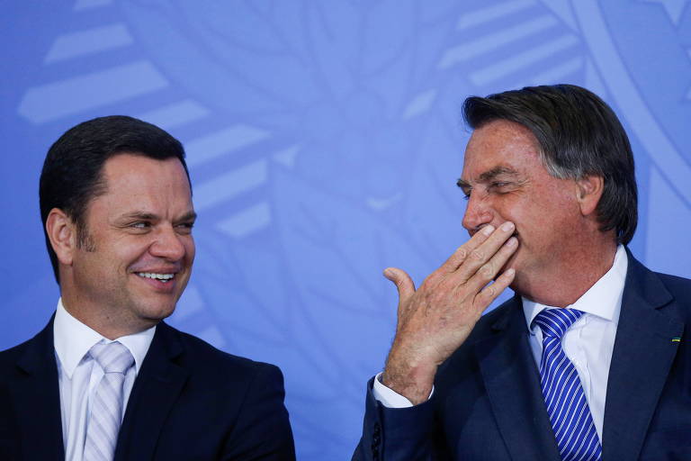 Anderson Torres e Bolsonaro sorriem um para o outro, lado a lado. Bolsonaro está cobrindo a boca com a mão
