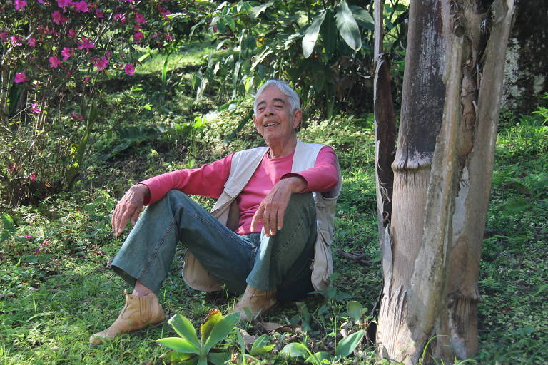 Leonardo Fróes é um homem magro, sorridente, de cabelos totalmente brancos; veste uma camiseta rosa e está em meio à vegetação