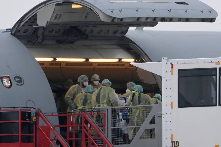 Médicos totalmente cobertos de branco, com máscaras e viseiras, transportam maca para dentro de um avião Airbus A310-900