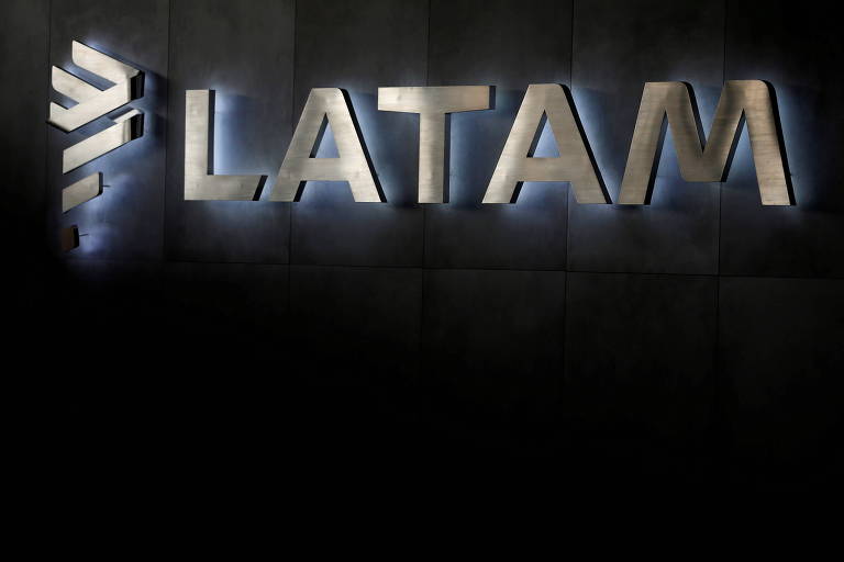 Logomarca Latam em prata, com luz na base de cada uma das letras, sobre uma parede preta