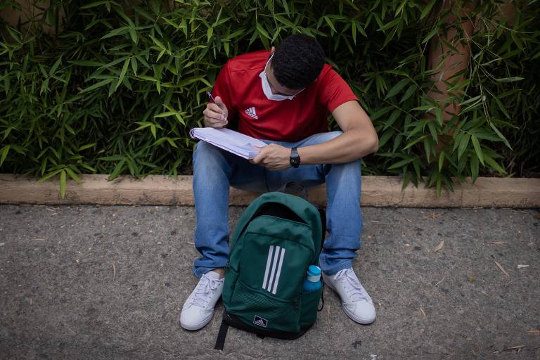 Um jovem sentado no chão, usando uma camiseta vermelha e um boné escuro, escreve em um caderno. Ele está cercado por plantas verdes e tem uma mochila verde ao seu lado. 