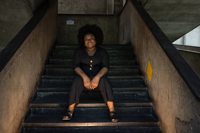 Negra, com cabelo natural, Loise Lorene, mestranda na UERJ que foi cotista no curso de psicologia da universidade, diante de escadaria no campus