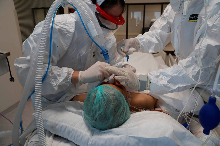 Dois médicos todos cobertos de branco, um de cada lado da maca, aplicam tubos nas vias respiratórias de um paciente deitado, de touca verde