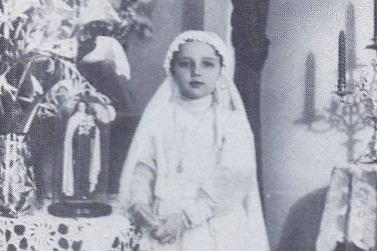 Imagem em preto e branco mostra criança com vestimenta branca. Ao seu lado, há a imagem de uma santa sobre uma mesa.