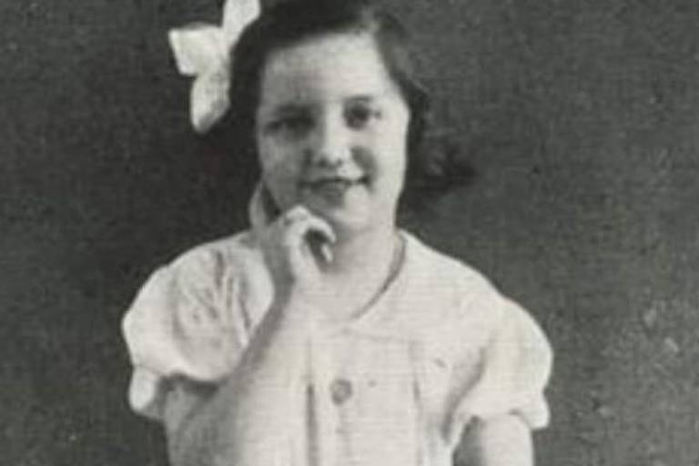 Imagem em preto e branco mostra uma garotinha sorrindo e com a mão apoiada no rosto próxima ao queixo