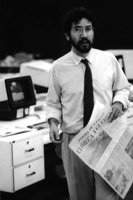 retrato em preto e branco de homem vestido com camisa, calça social e gravata está em pé em um meio a mesas e computadores segurando um jornal com a manchete "começa a guerra" 