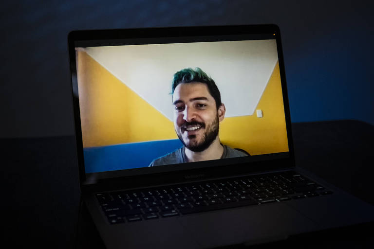 Eduardo aparece sorridente numa tela de computador do tipo notebook, em frente a um fundo de parede pintada em branco e amarelo. 