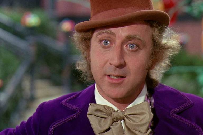 Foto mostra Gene Wilder interpretando Willy Wonka em A Fantástica Fábrica de Chocolate. Ele é um homem branco, de cabelos loiros e olhos azuis, e veste um colete roxo, gravata borboleta bege e uma cartola marrom clara