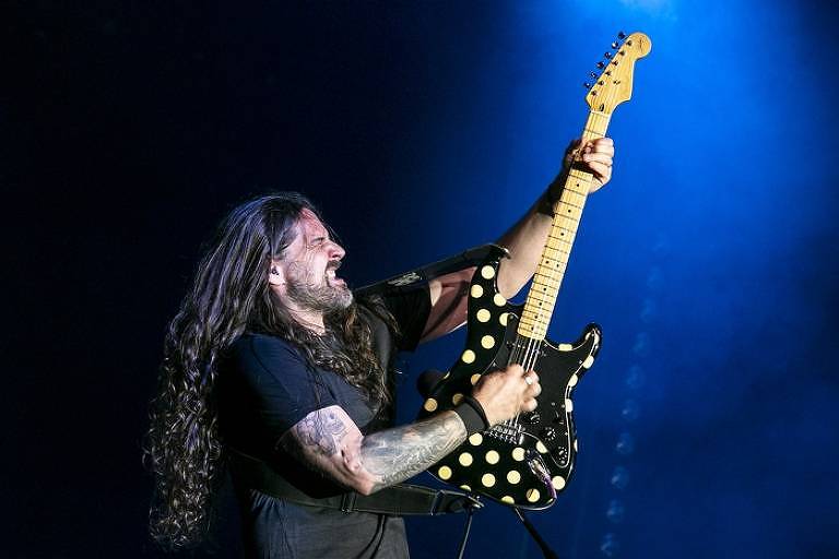 Com cabelos longos e pretos, e o braço direito tatuado, o guitarrista Andreas Kisser empunha a guitarra preta com bolas amarelas, levantando-a para cima
