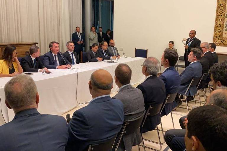Bolsonaro, Mendonça e outras pessoas sentados à mesa. Diante dele, duas fileiras de homens sentados, usando terno
