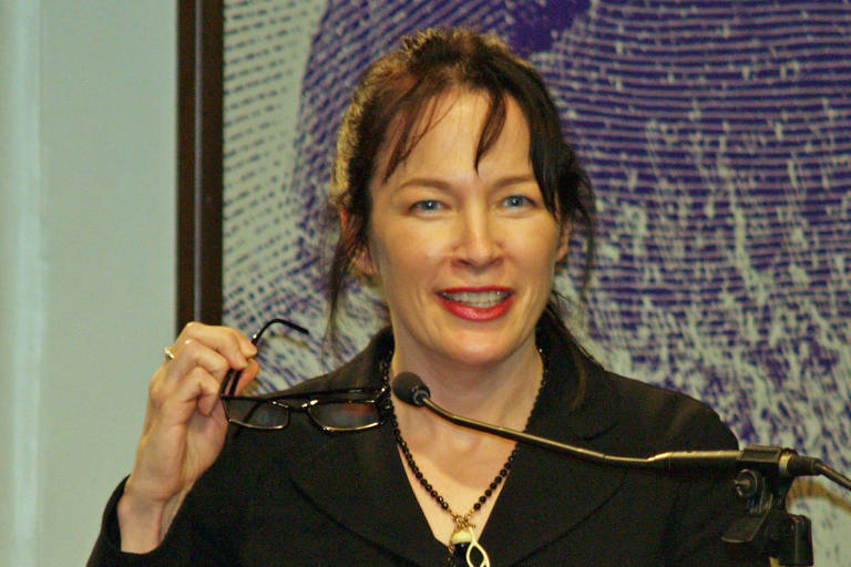 fotografia de uma mulher branca diante de um microfone segurando seus óculos