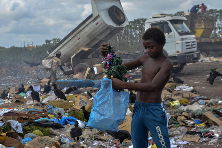 Um menino no meio de um lixão segura uma árvore de natal que encontrou em uma sacola plástica