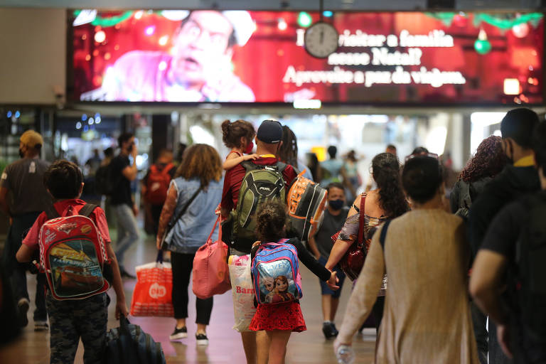 pessoas de costas no aeroporto caminham com malas, família em destaque tem homem segurando criança de colo dando a mão a outra, que anda e carrega mochila nas costas