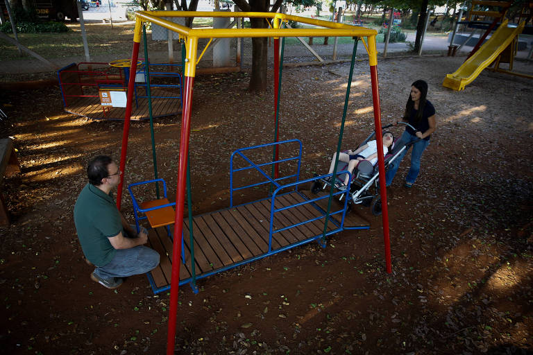 Imagem destaca um balanço acessível para crianças com deficiência em uma praça; a estrutura é colorida e contém um piso de madeira: de um lado, há o espaço para uma cadeira de rodas, do outro, um assento para ser utilizado por crianças sem deficiência, para que brinquem juntas