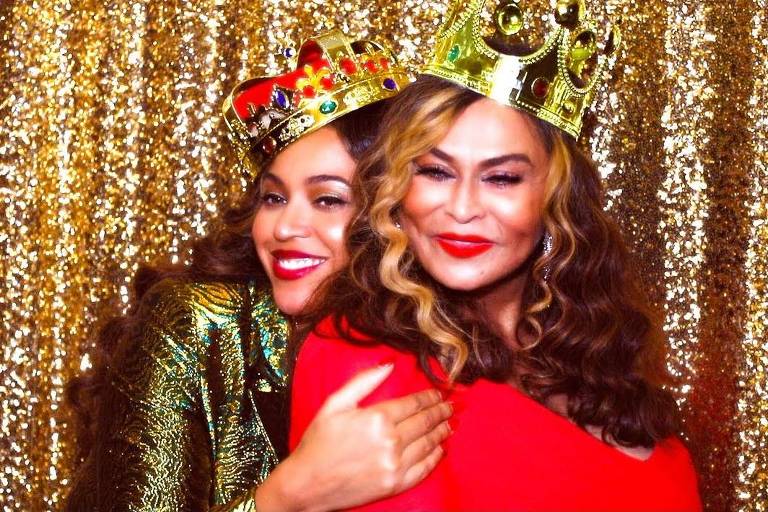 Beyoncé e sua mãe, Tina Knowles, com vestidos de festa e coroas de papel na cabeça. Ambas são mulheres negras, de cabelos longos e castanhos, e estão abraçadas em frente a cortina dourada