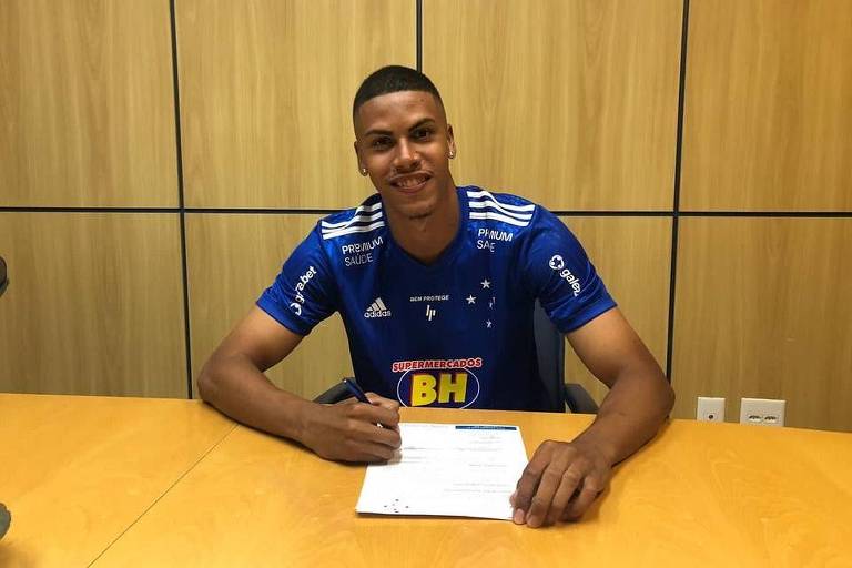 Zagueiro Paulo Eduardo, homem negro de cabelo curto, sorridente, vestindo a camisa azul marinho do time Cruzeiro enquanto assina o contrato com o clube
