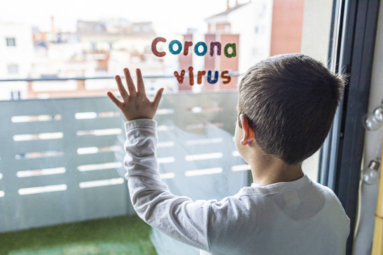 De costas, um garotinho branco, com cabelos curtos e castanhos aparentando ter cinco anos está em pé com a palma da mão esquerda encostada no vidro da janela. Ao lado da mão, a palavra coronavírus.