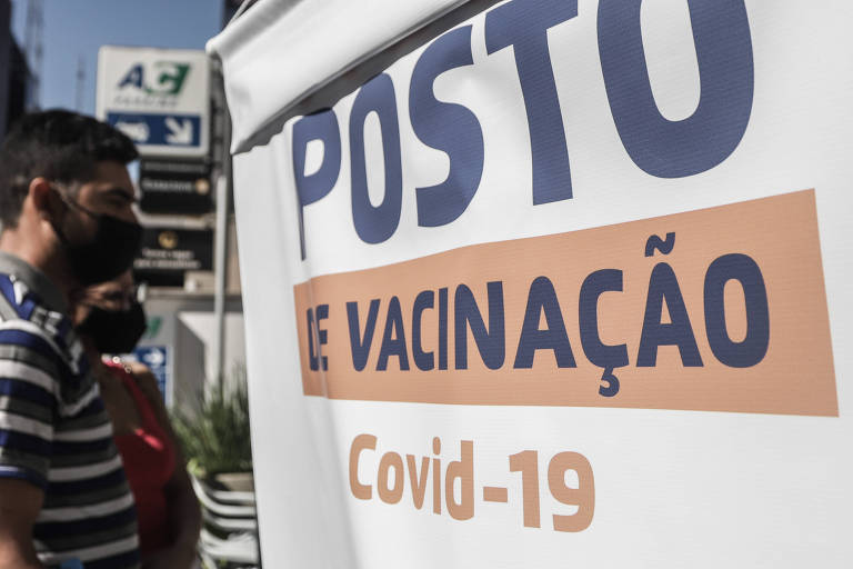Pessoas olham banner onde se lê "Posto de vacinação Covid-19" em dia de sol