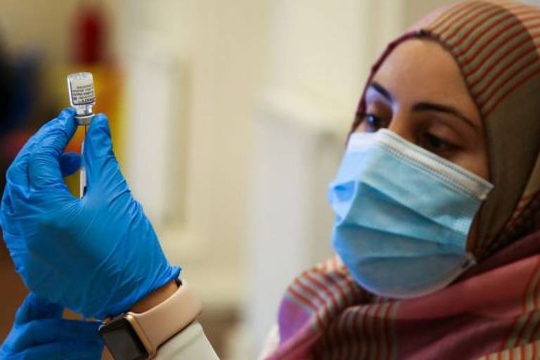 Imagem em primeiro plano mostra mulher de máscara e luvas segurando uma seringa espetada em uma ampola de vacina