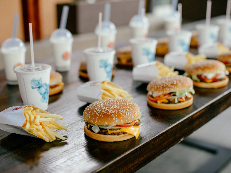 Uma mesa de madeira apresenta uma variedade de alimentos de fast food， incluindo hambúrgueres com gergelim， porções de batatas fritas e copos de bebidas. Ao fundo， há copos de milkshake. Os hambúrgueres estão dispostos em linha， com ingredientes visíveis como queijo， alface e tomate.