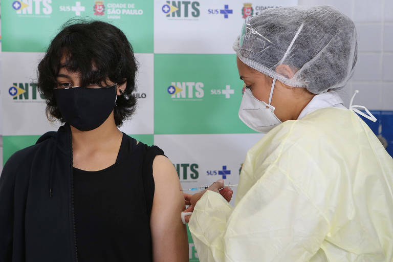 Agente de saúde, de avental, touca e máscara, aplica vacina em mulher branca, de blusa e máscara pretas