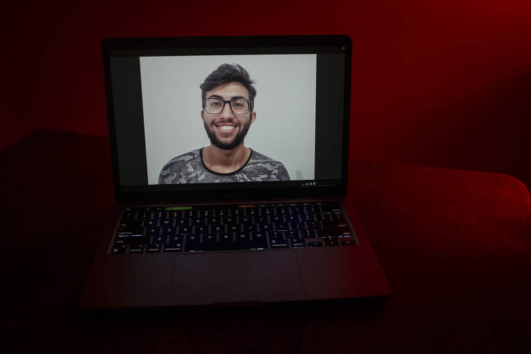 Imagem de Lucas sorrindo aparece na tela de um notebook; ele é branco, usa óculos e tem barba; no entorno, o ambiente está escuro