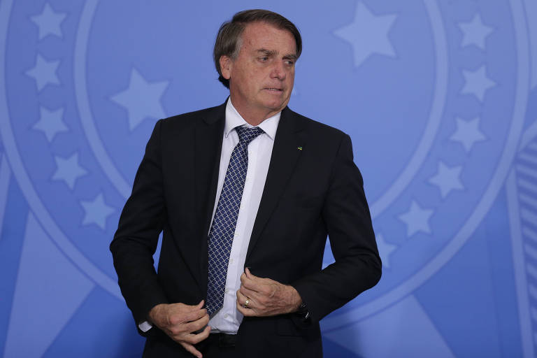 Bolsonaro, de paletó escuro, camisa branca e gravata, está em pé; ao fundo, painel azul com estrelas