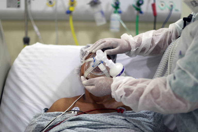 Foto mostra médico, com máscara, luvas e EPI ajustando a máscara de oxigênio de um homem, que está deitado numa maca. 