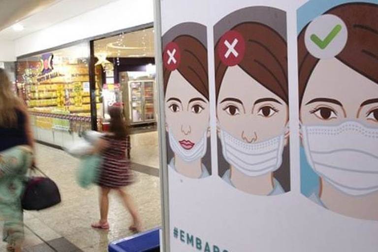 Em destaque, cartaz mostra ilustração de como se deve usar a máscara. Ao fundo, há uma mulher e uma criança caminhando em um corredor.