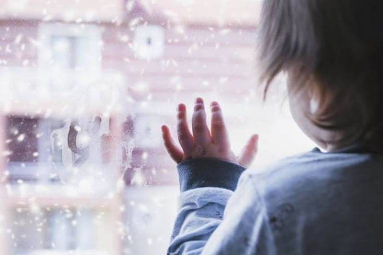 De costas, uma criança está com uma das mãos apoiada em uma janela de vidro