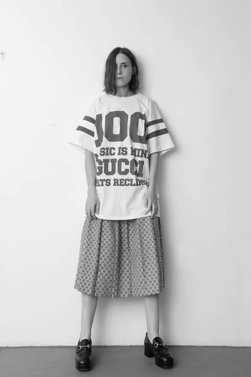 retrato em PB de mulher com camiseta grande, saias e mocassins de salto diante de parede branca