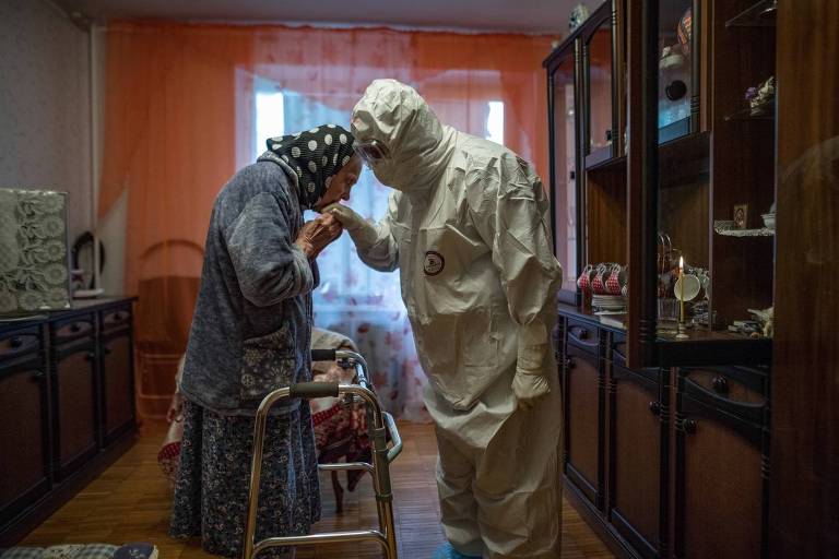 Mulher idosa aparece beijando a mão de um enfermeiro. O profissional de saúde usa traje de segurança, que cobre todo o corpo. Os dois se encontram em uma cozinha cheia de móveis de madeira.