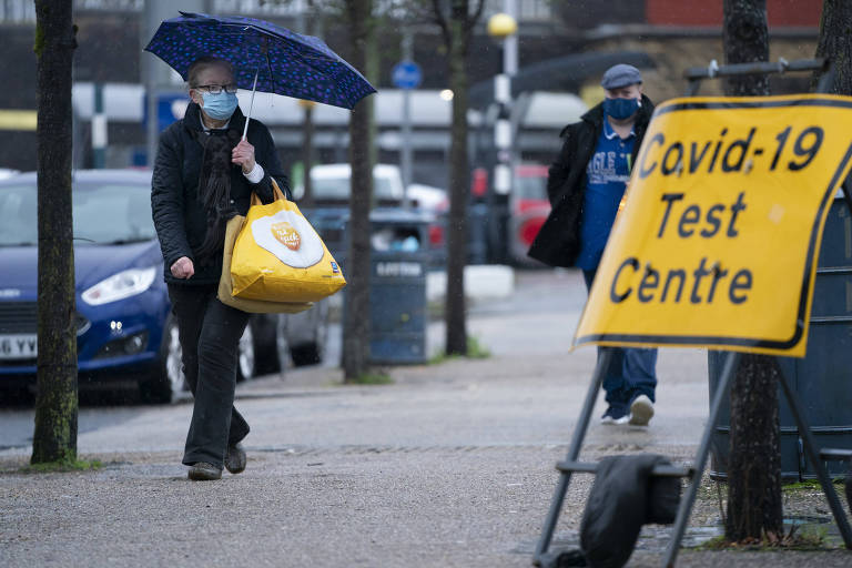Homem de máscara segurando um guarda-chuvae  sacolas caminha em uma rua. No canto direito da imagem, há uma placa escrito: Covid-19 Teste Centre